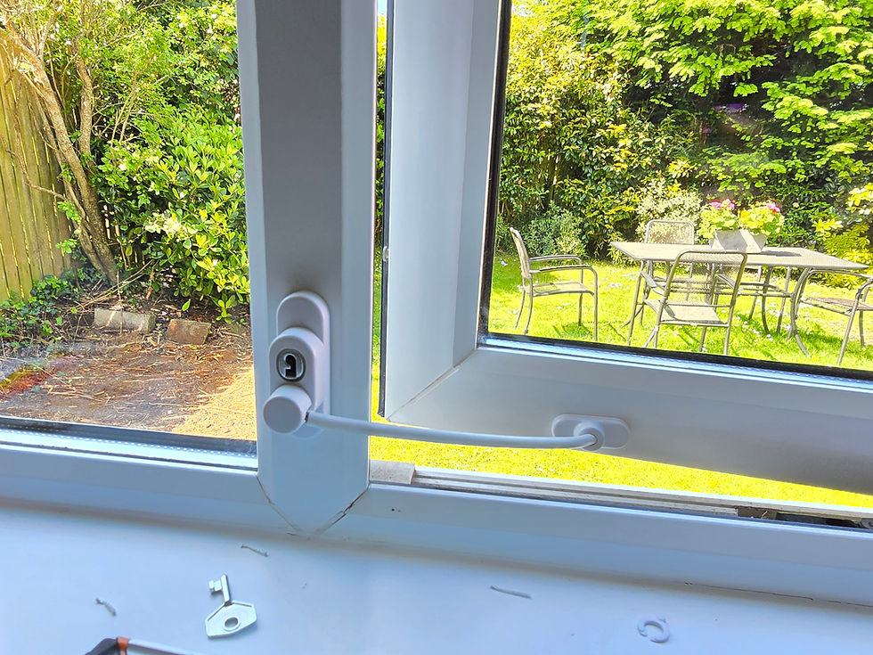 Open window with a white lock, key on the sill, view of a sunny garden with green plants and patio furniture visible outside.