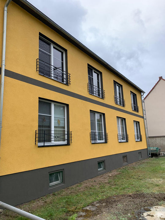 Renoviertes Mehrfamilienhaus mit moderner Putzfassade in Gelb und dunklen Akzenten. Neue Fenster mit stilvollen französischen Balkonen sorgen für ein zeitgemäßes Erscheinungsbild. Solide Bauweise mit langlebiger Fassadengestaltung und energieeffizienter Dämmung. Begrünter Außenbereich mit gepflegtem Fundament.