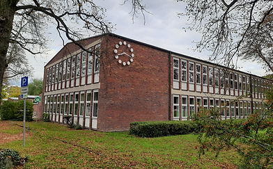 Der Kopfbau mit großer Uhr in der linken Ecke, umgeben von Hecken und Wiese.