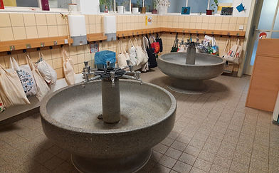 Gefliester Waschraum mit Rundwaschbecken aus Stein mit mehreren Wasserhähnen, im Hintergrund hängen Taschen an der Garderobe.