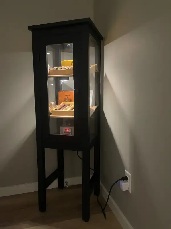 Tall and narrow black wooden cigar humidor cabinet with elevated legs, illuminated by spotlight, featuring glass door and several shelves of assorted cigars, with digital hygrometer reading, in a dimly lit room