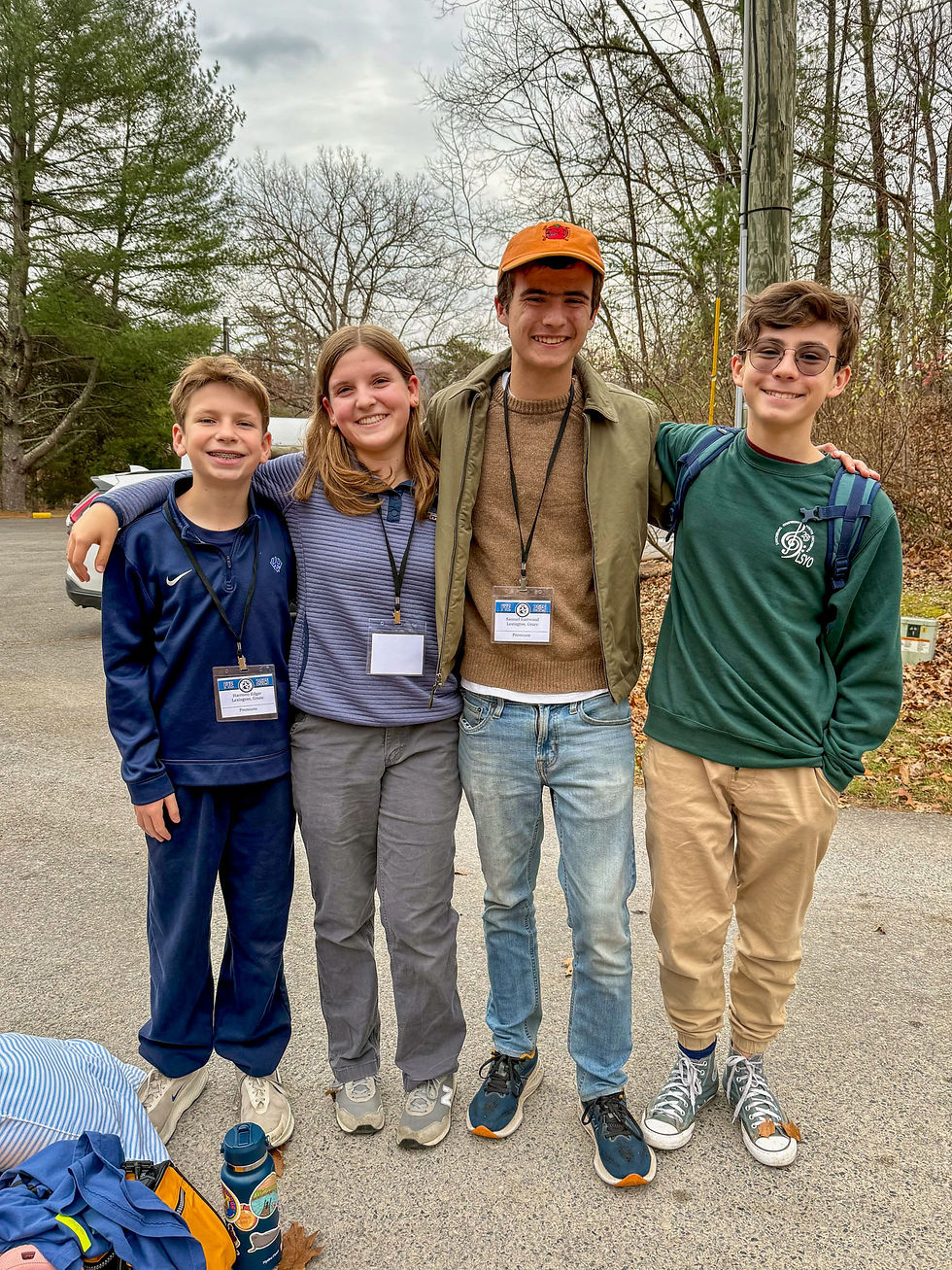 Four smiling teens stand together outdoors on an overcast fall day, arms around each other. They are wearing casual clothes and retreat name badges on lanyards. Bare trees and a few evergreens are visible behind them along with a utility pole and parked cars. A duffel bag and water bottle rest on the ground in front of them.