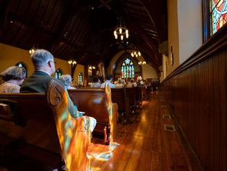 Congregants sit in wooden pews during a worship service inside Grace Episcopal Church, with sunlight from stained-glass windows casting colorful reflections across the pews and floor.