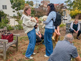 A group of people gather outdoors in a small garden area, chatting and holding fluffy white puppies. Two women in sweaters and jeans stand facing each other, each holding a puppy. Others sit on benches nearby, smiling and talking. Autumn leaves are scattered on the ground, and a church building with a sign reading “Grace” is visible in the background.