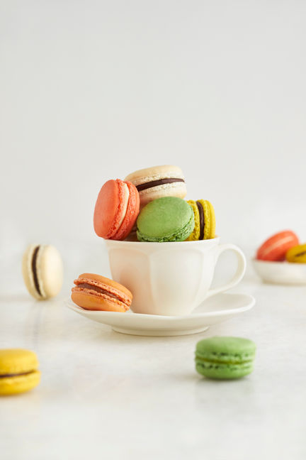 A set of colourful macarons of assorted flavour fill a white teacup. There are a few more strewn around on the bright white setup.