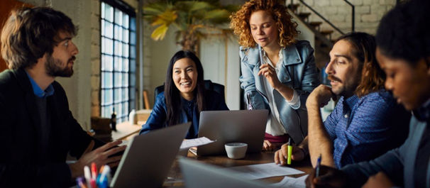 Group of people collaborating in a modern office with laptops and notebooks. Bright mood with natural light and green plant decor.