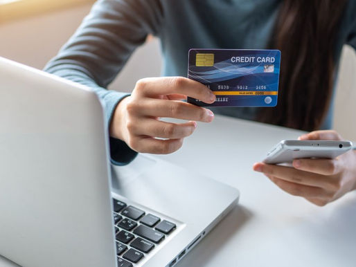 Person holds a credit card in one hand and a smartphone in the other, next to a laptop on a desk—highlighting the convenience of digital payments and the hidden impact of processing fees. Bright background with focus on the card.