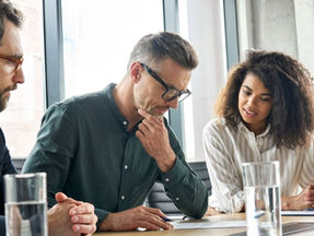 Three people in a meeting room reviewing documents, representing a professional services team. Large windows in the background with natural light, glasses of water on the table, and a serious, collaborative mood.