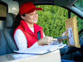 Smiling delivery driver in a red cap scanning a package inside a van, representing real-world logistics supported by a unified ERP for CPG brands, with parcels, clipboard, and green foliage in view.