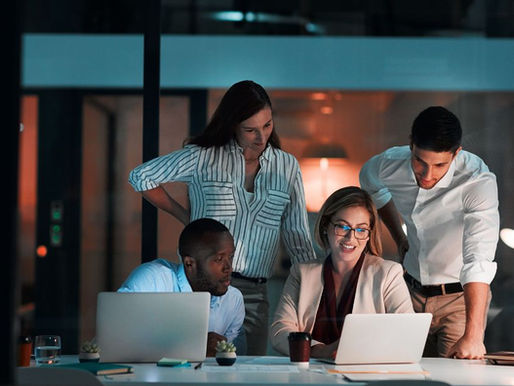 Four colleagues gather around a laptop in a warmly lit office at night, discussing how to boost productivity using Microsoft Copilot. The collaborative mood highlights teamwork and innovation.
