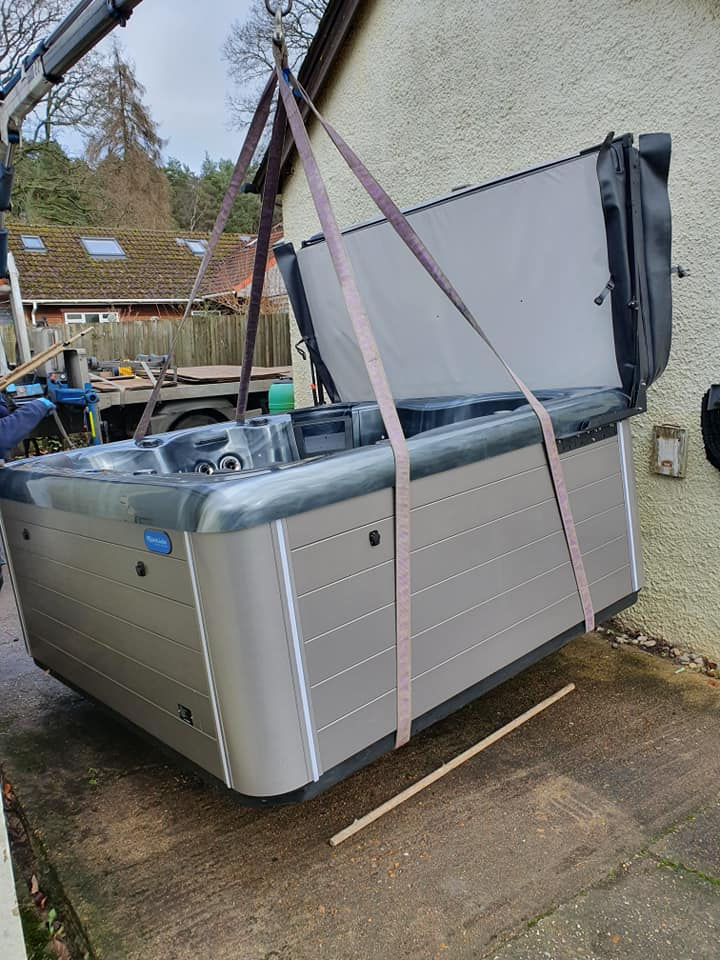A hot tub is being carefully lifted with a crane to be installed beside a house, against the backdrop of a garden and nearby homes.
