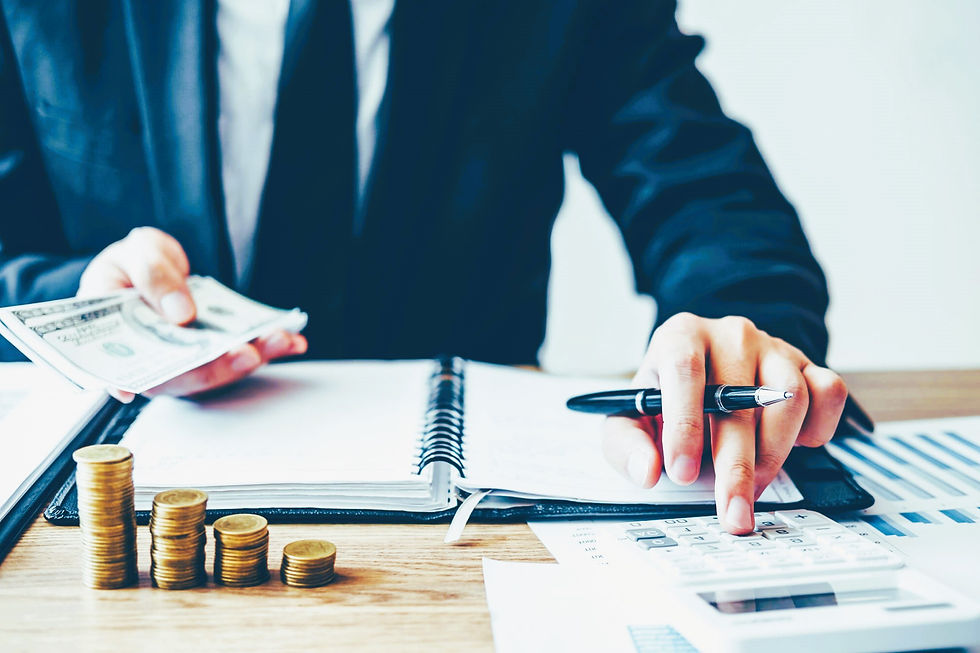 Person in suit calculates with a pen while holding cash, notebook open. Coins stacked on desk, charts and calculator visible. Business setting.