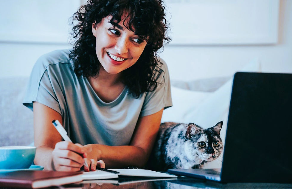 Woman smiling while writing in notebook near a laptop. A cat sits beside her on a couch. Blue cup and relaxed atmosphere.