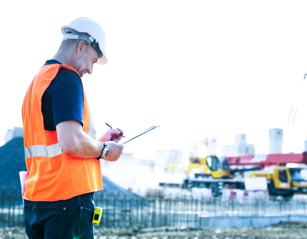 Construction worker in an orange vest and hard hat writes on a clipboard at a site. Background shows cranes and equipment under bright sky.