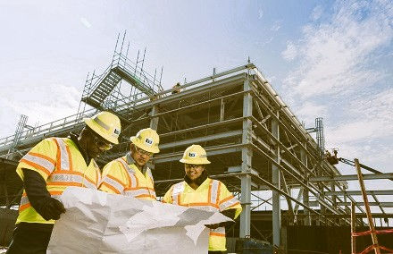 Construction workers in yellow vests examine blueprints at a building site with steel frameworks under a blue sky. Safety and teamwork evident.