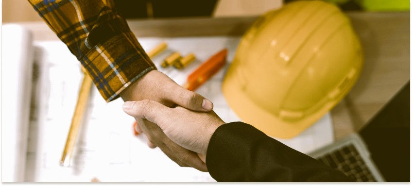 Two people shaking hands over architectural plans, a yellow hard hat and tools in the background, suggesting a business agreement.