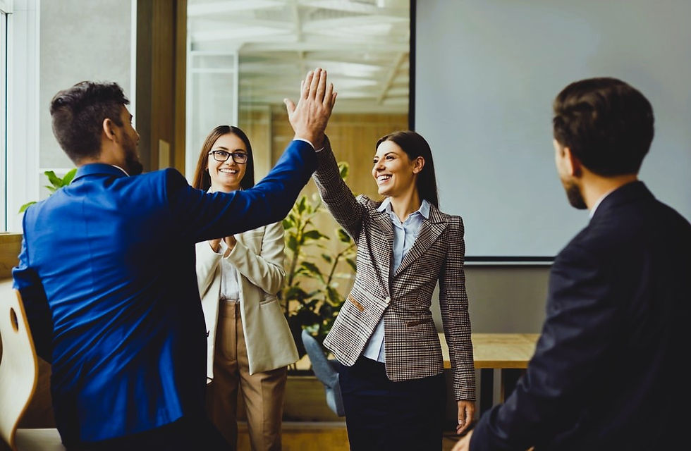 Four people in an office celebrate a success; two high-five, others clap and smile. Bright room with plants, creating a cheerful mood.