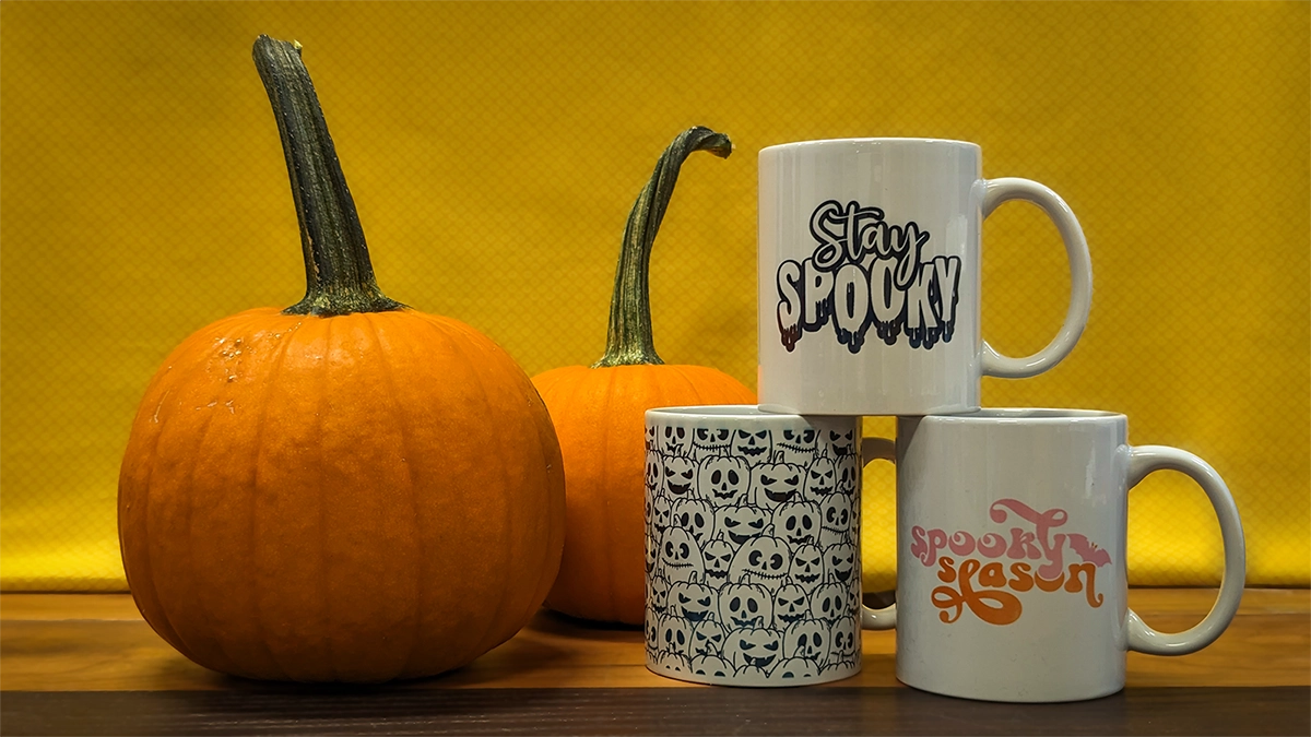 Three mugs sit to the right of pumpkins. The top mug says Stay Spooky, the bottom right says Spooky Season, the bottom left is covered in jack o lanterns