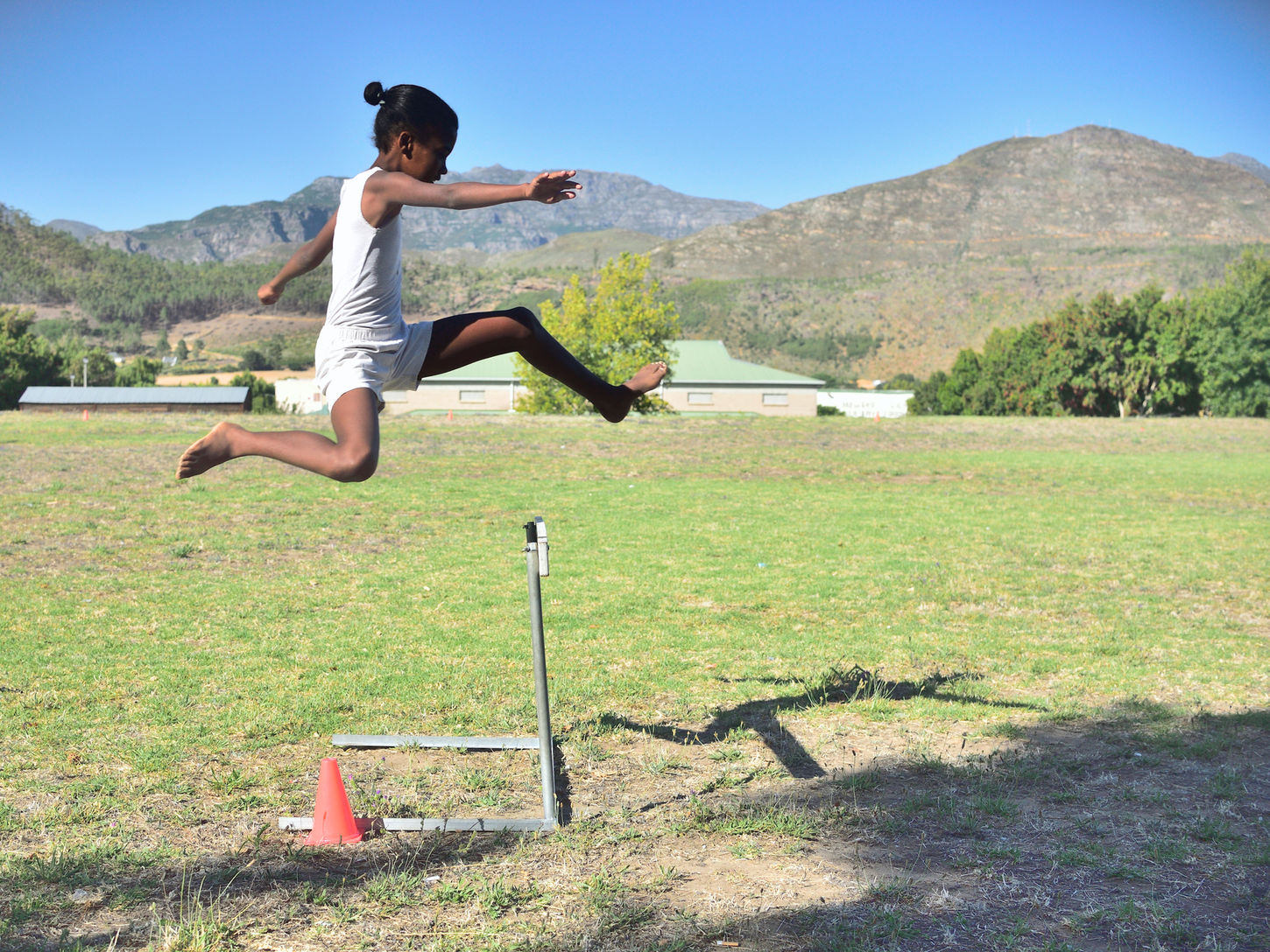 A barefoot girl leaps over a low training obstacle on a grassy field. Her shadow stretches across the ground, with mountains and a clear blue sky in the background.