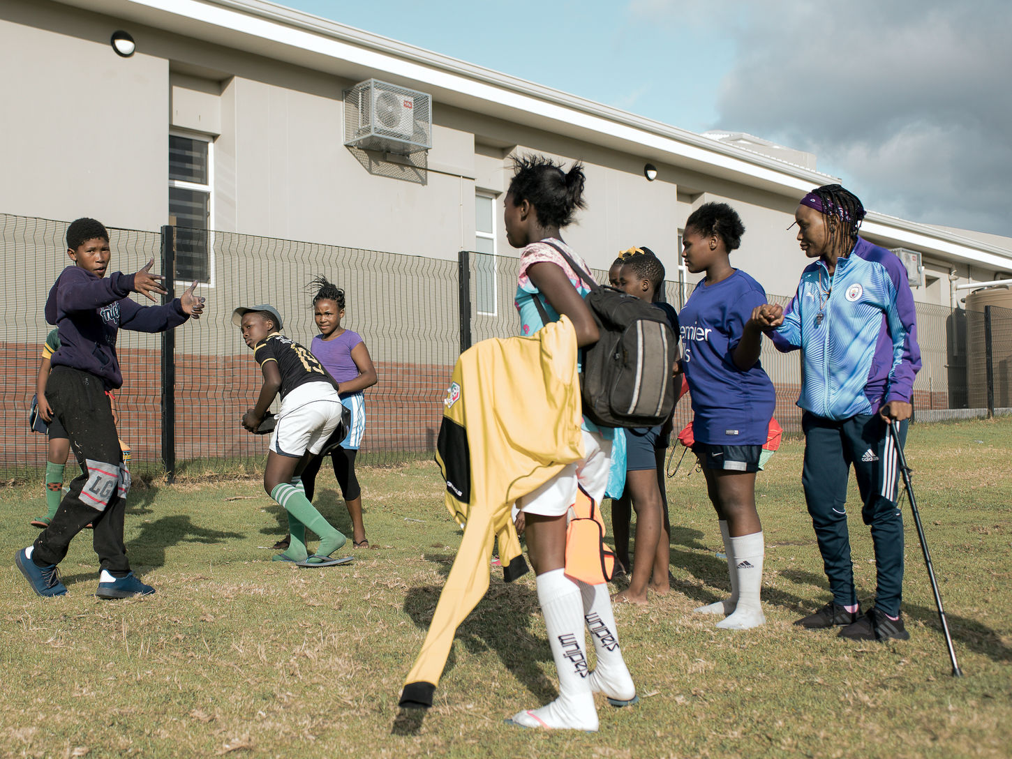 Teenagers gather outside a sports field while a boy gestures toward a group of girls during an animated conversation. They wear sports clothing and carry training gear.