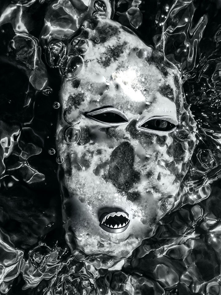 Black-and-white image of a weathered African mask surrounded by flowing water and air bubbles, with a stylised mouth suggesting a hidden presence.