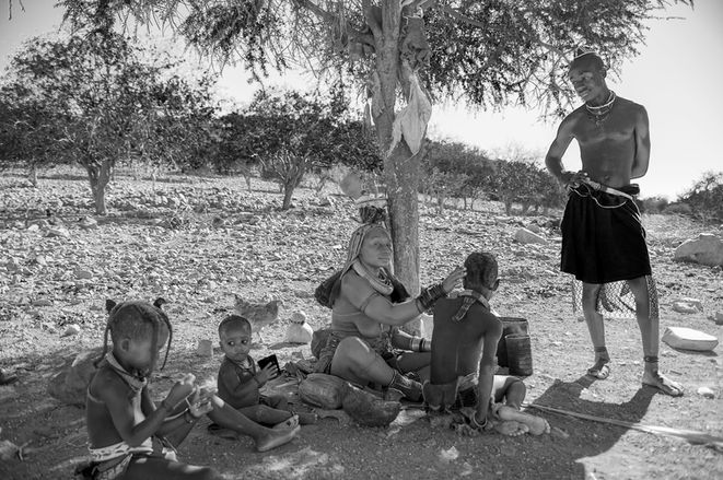 : Himba family gathered beneath the shade of a tree in northern Namibia. A mother touches a child's head while other children sit nearby and a young man stands beside the group.

