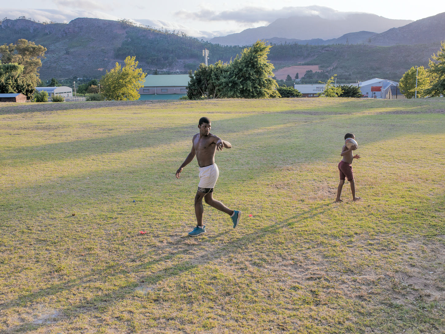 A young father and his son cross a wide grassy field at sunset. The father gestures while the child walks nearby carrying a rugby ball over his shoulder, with long shadows, mountains and scattered buildings in the background.