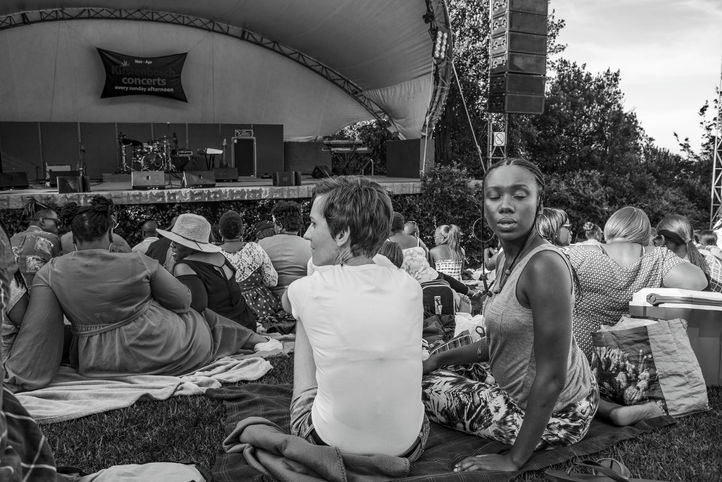 A young woman sits among spectators and turns toward the camera with her eyes closed, while people face an outdoor stage in the background.