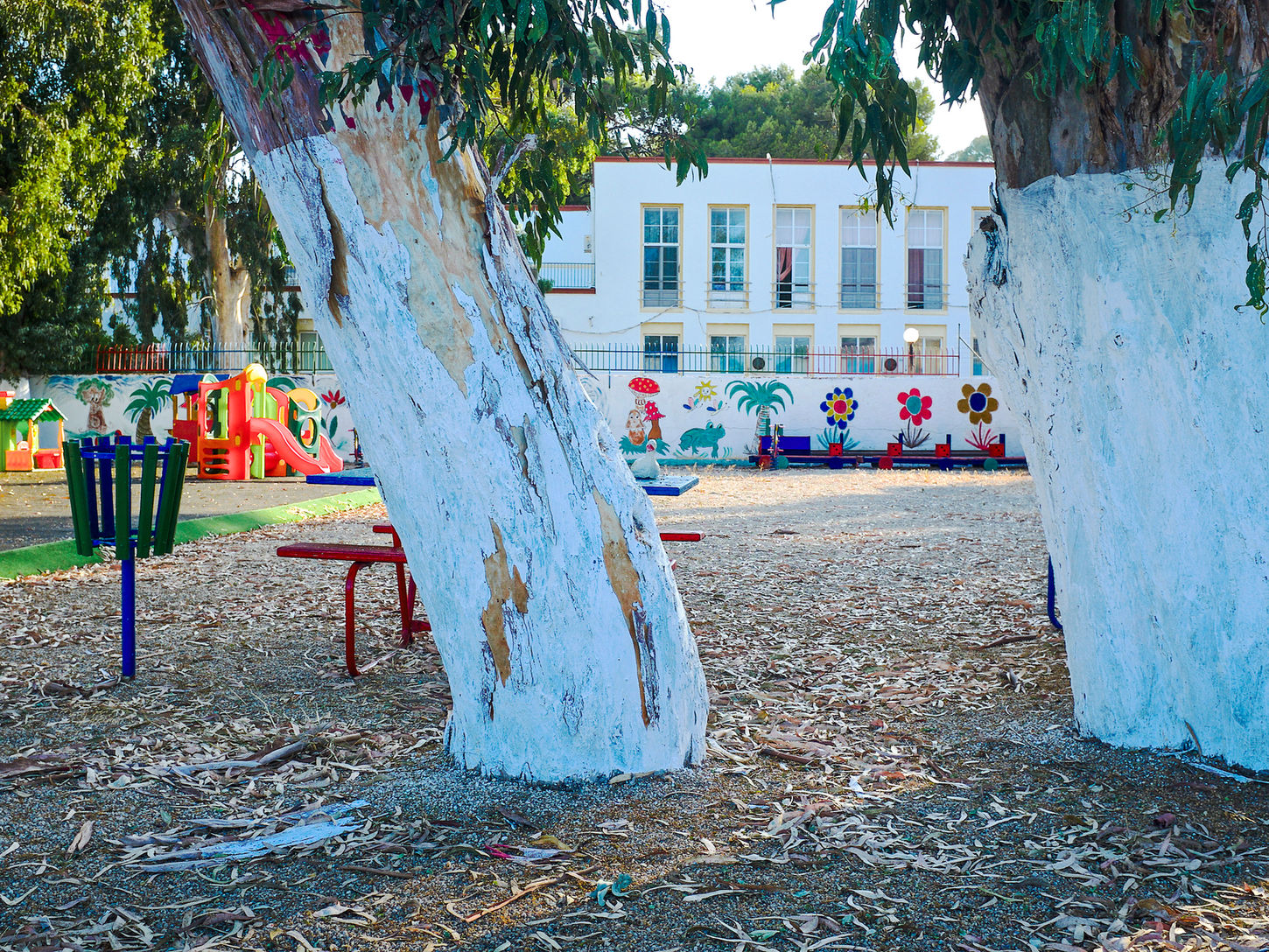 An empty playground is framed by two white-painted tree trunks. Colourful play structures, painted murals and a white building appear beyond the trees, surrounded by green foliage.