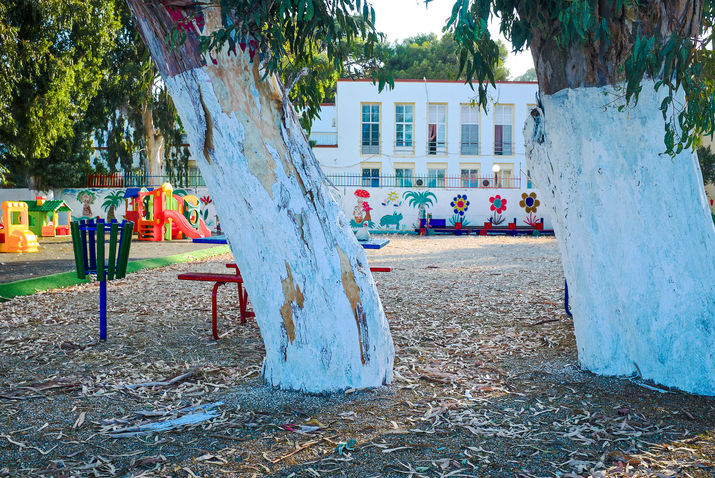 An empty playground is framed by two white-painted tree trunks. Colourful play structures, painted murals and a white building appear beyond the trees, surrounded by green foliage.

