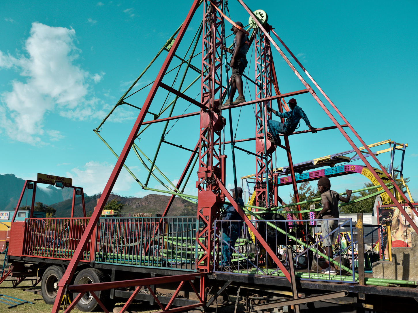 Fairground workers climb the metal structure of a ride during its assembly as others work below. The geometric framework stands against the blue sky, with mountains and other attractions beyond.