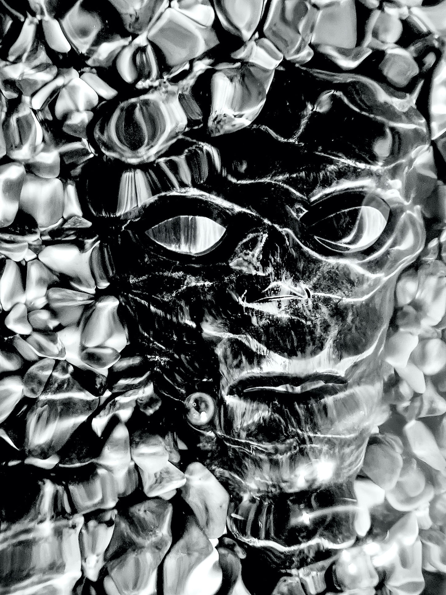 Black-and-white image of an African mask emerging from smooth seashore stones beneath moving water, with shimmering reflections across its surface.