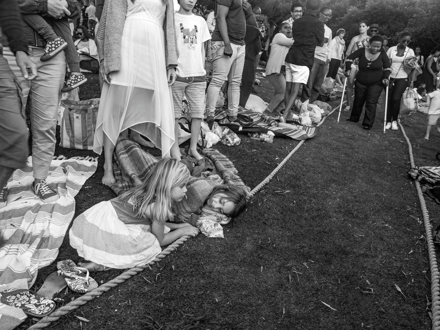 A young girl kneels beside another girl lying on the grass in the foreground, while a woman using crutches walks toward the camera with a younger woman along a central aisle at an outdoor event.