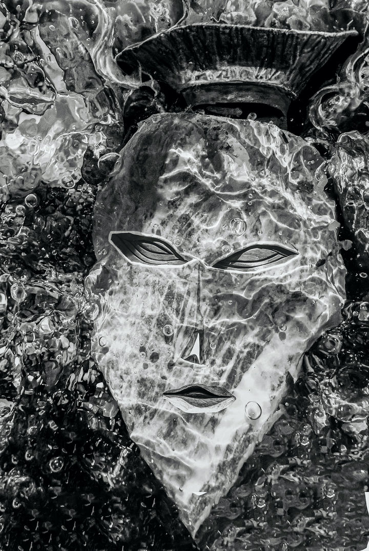 Black-and-white image of an African mask partially veiled by bright reflections created by moving seawater over smooth stones.