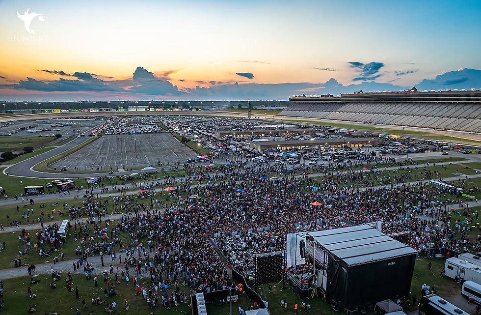 Crowd gathered at a race track during sunset, with a stage in the foreground. Vibrant sky above, creating a festive atmosphere.