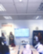 Three women present in a classroom with a map on a screen. Students listen attentively. Background shows "asphaleia training" text.