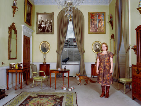 Woman in floral dress standing in a yellow 18th-century drawing room with portrait paintings, a fox, and a crow figure on the floor