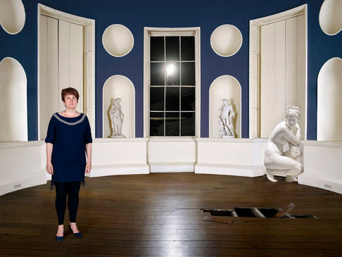 Woman in navy dress standing in an oval blue room with classical statues in wall niches and an open trapdoor in the floorboards