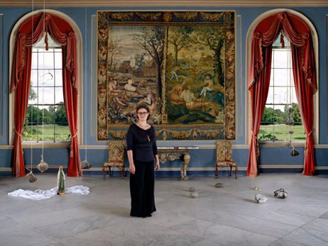 Woman in black dress standing in a blue tapestried room with objects suspended above the floor and a view over green fields through arched windows