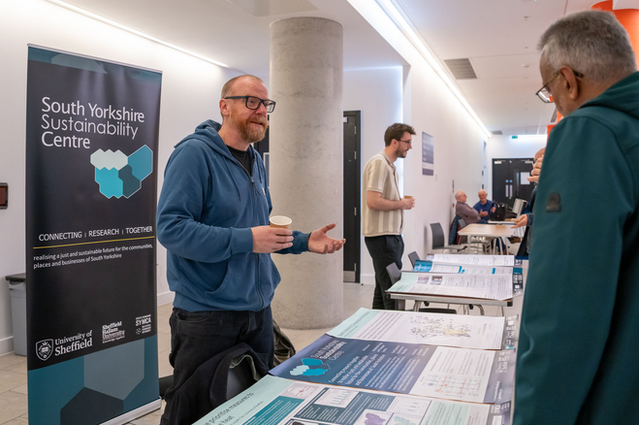 A person wearing a blue hoody and black glasses addressing another person. To the right of them is a banner with the branding of South Yorkshire Sustainability Centre. In front of the person is a table full of posters.