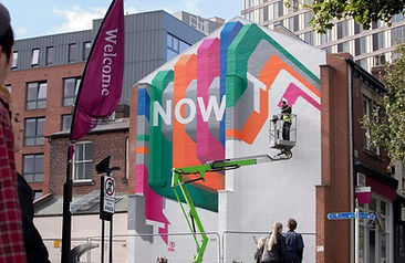 A photo showing a big mural on the gable end of a house, half completed. Visible are the words "Now" and the leter T, and a person on a crane continuing to paint the rest of the mural in a colourful, abstract design