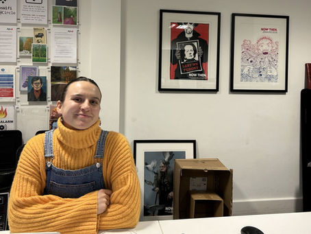 A photo of a woman wearing dungarees and an orange jumper sitting at a desk in an office, smiling