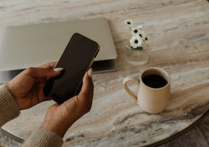 Person holding a smartphone over a marble table with a laptop, a cup of coffee, and a small vase of white flowers, creating a calm and minimal workspace scene.