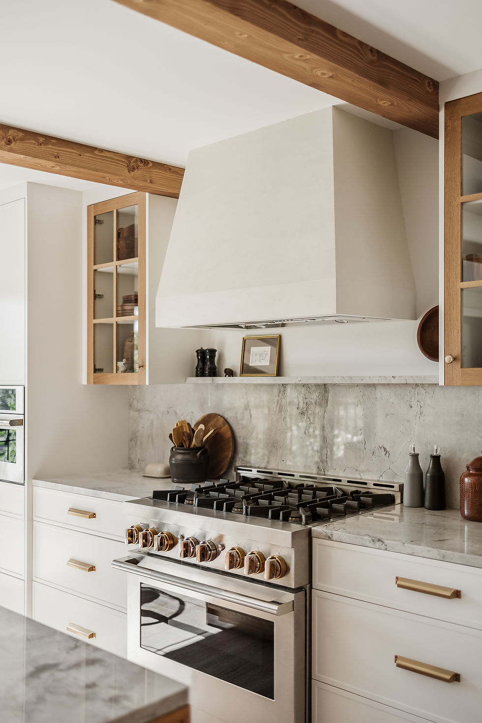Light, modern farmhouse kitchen with exposed wood ceiling beams, white cabinetry, marble backsplash, and a stainless steel range with brass hardware.