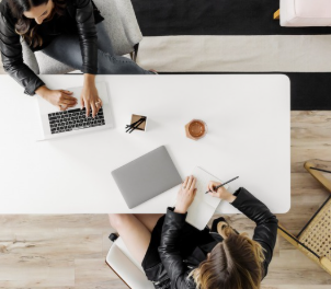 Overhead view of two women working at a white desk with a laptop, notebook, and coffee in a modern office.