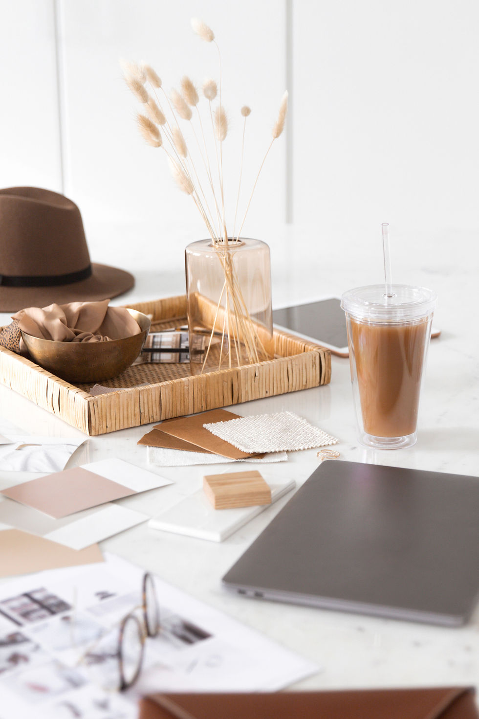 Styled creative workspace with fabric swatches, neutral color samples, a laptop, iced coffee, and decorative accents arranged on a bright white surface.