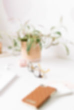 A pair of round eyeglasses rests on a white desk alongside a potted plant, pink wireless earbuds, a notepad, and a brown polka-dotted phone case, creating a soft and minimal workspace scene.