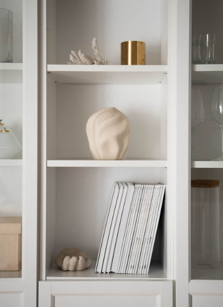 Minimal white built-in shelving styled with neutral decor, including stacked books, a sculptural ceramic object, coral decor, and a brass accent piece.
