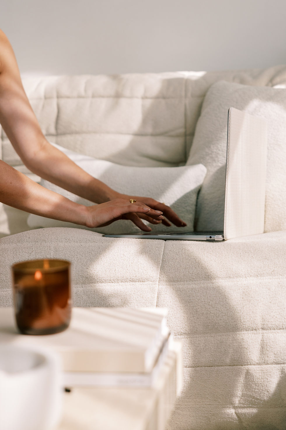 Person working on a laptop while sitting on a soft cream-colored sofa, with a lit candle and stacked books on a nearby table.