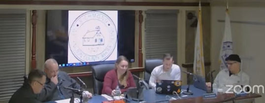 Town council members gather for a meeting, discussing community affairs with a town seal displayed on a screen behind them as the session is streamed live on Zoom.